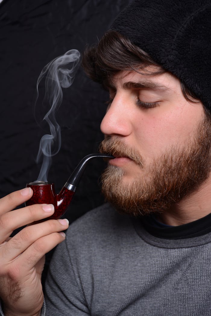 Close-up portrait of a bearded man thoughtfully smoking a pipe indoors.