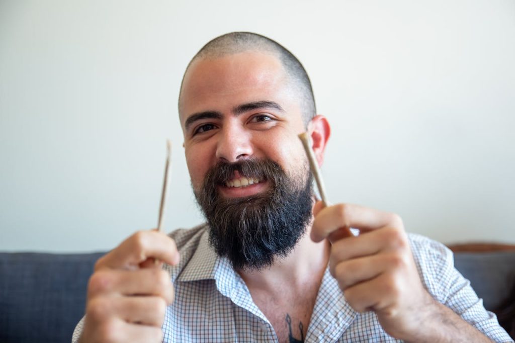 A cheerful bearded man with a checkered shirt holding two cannabis joints indoors.