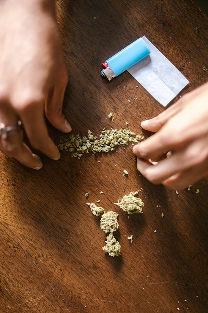Hands rolling a cannabis joint with a lighter and paper on a wooden surface.